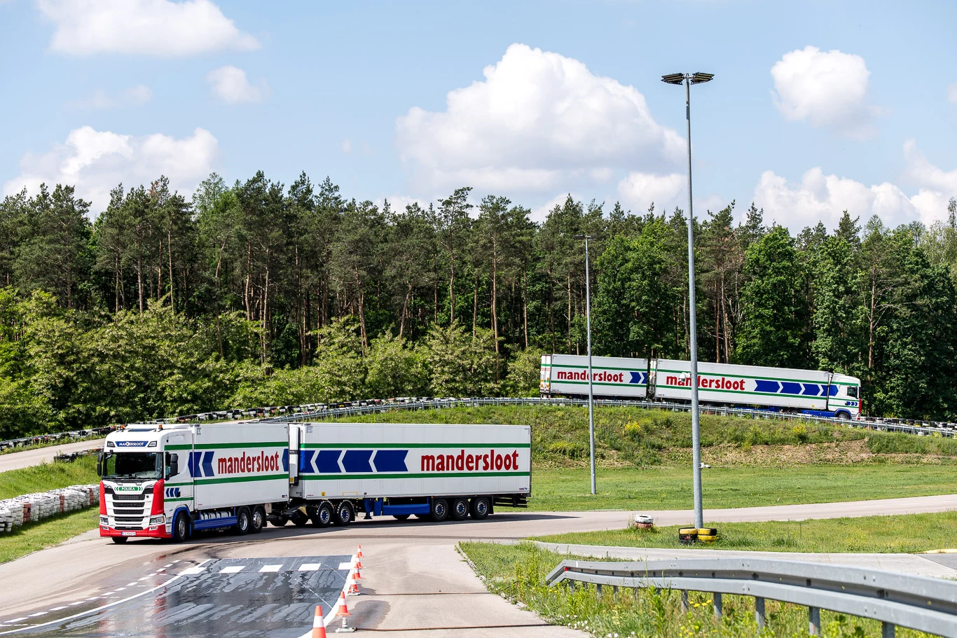 Długi zestaw ciężarowy Mandersloot manewruje na krętej trasie w otoczeniu zielonego lasu pod jasnym niebem.