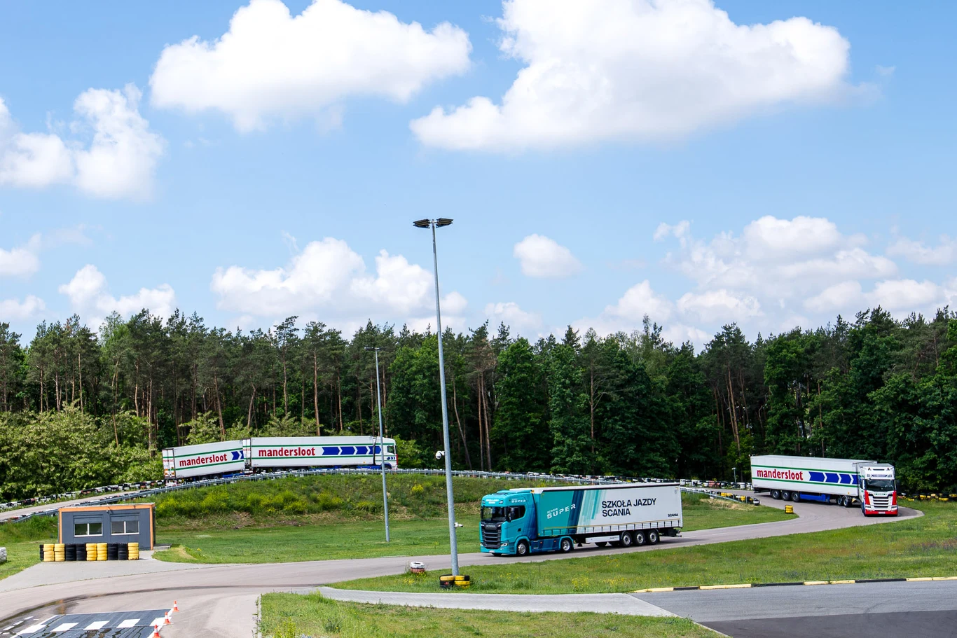 Ciągniki siodłowe z naczepami poruszające się po torze manewrowym otoczonym zielenią, z budynkiem technicznym w pobliżu oraz lasem w tle.