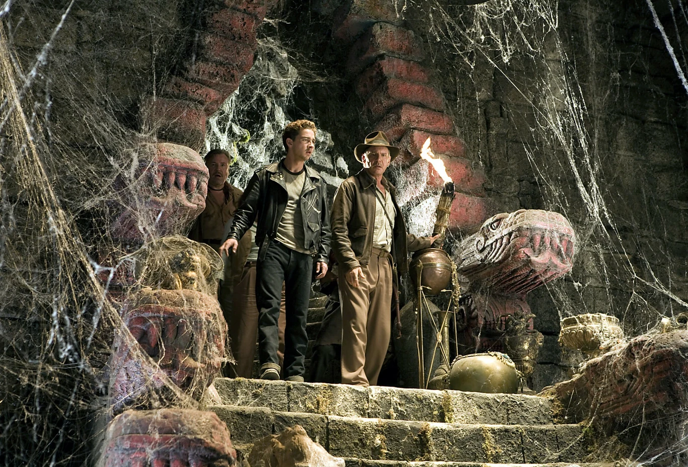 Shia LaBeouf i Harrison Ford w filmie "Indiana Jones i królestwo Kryształowej Czaszki" Trzech mężczyzn idzie po kamiennych schodach wewnątrz świątyni ozdobionej rzeźbami w kształcie węży, otoczonych pajęczynami i tajemniczym światłem pochodni.