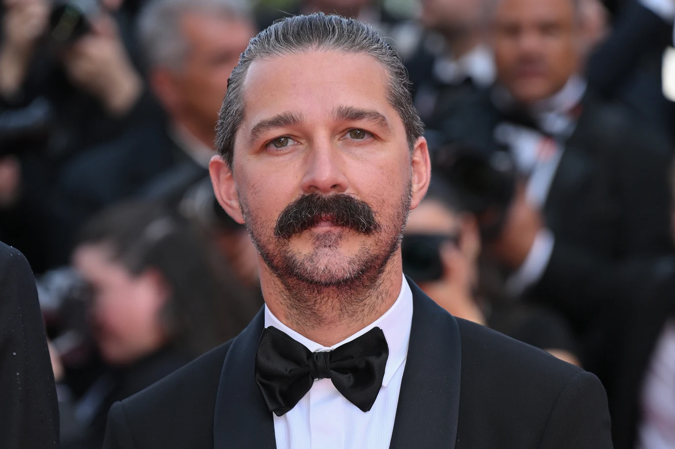 Shia LaBeouf w 2025 roku Mężczyzna z ciemnymi włosami zaczesanymi do tyłu i wyrazistym wąsem, ubrany w klasyczny smoking z białą koszulą i czarną muchą, na tle tłumu ludzi w eleganckich strojach.