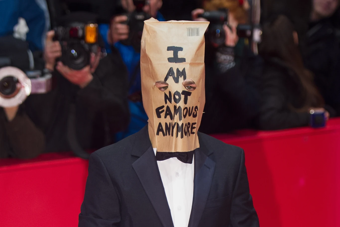 Shia LaBeouf w 2014 roku Osoba ubrana w elegancki garnitur z papierową torbą na głowie, na której widnieje napis „I am not famous anymore”, otoczona tłumem fotoreporterów z aparatami i fleszami.