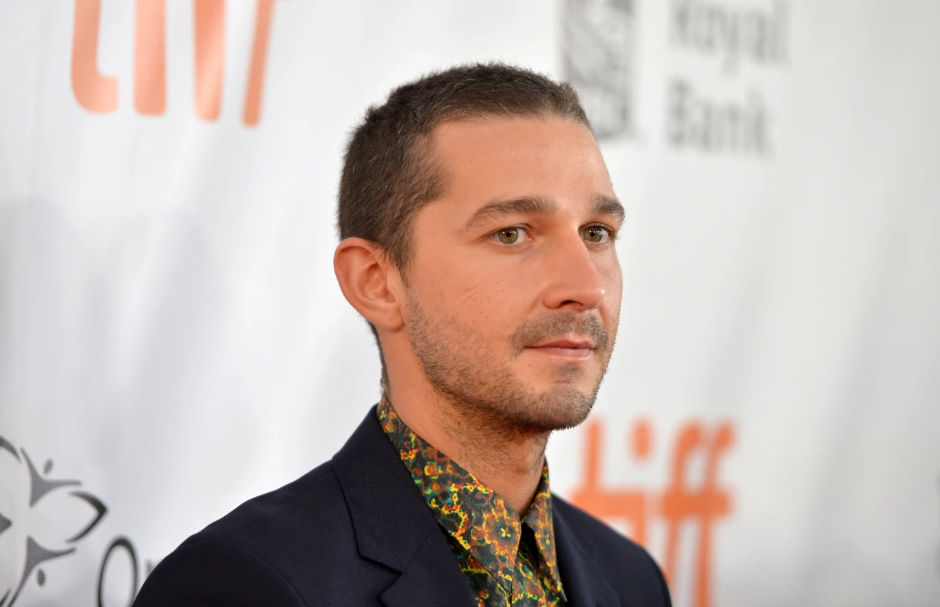 Shia LaBeouf w 2017 roku Mężczyzna w ciemnej marynarce i kolorowej koszuli stoi przed ścianką z logotypami, patrząc lekko w bok.