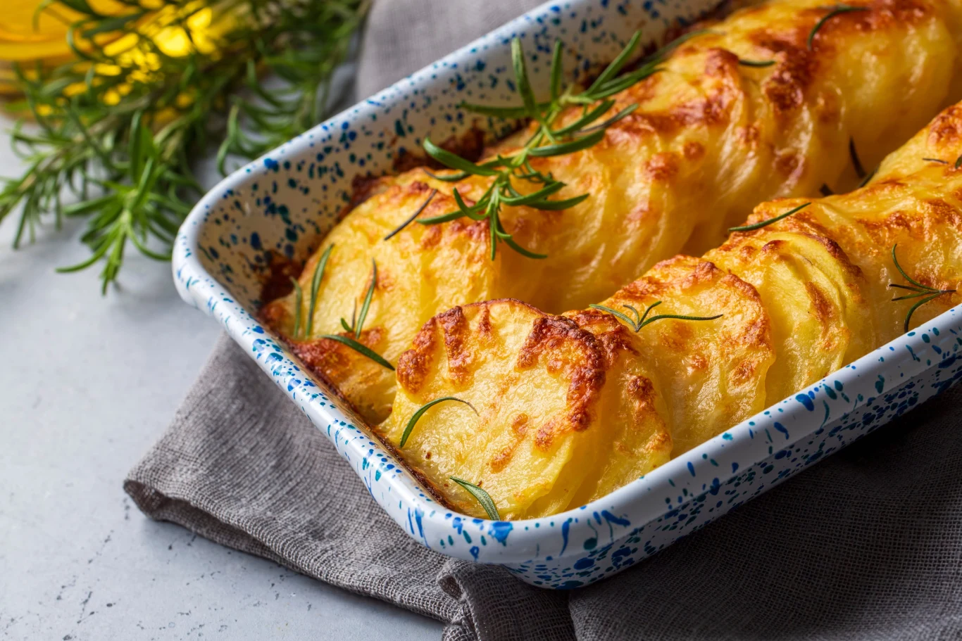Potato gratin - gratin (baked potatoes with cream and cheese) with rosemary and forks Zapiekane plastry ziemniaków z serem i śmietanką w ceramicznym naczyniu, udekorowane gałązkami rozmarynu