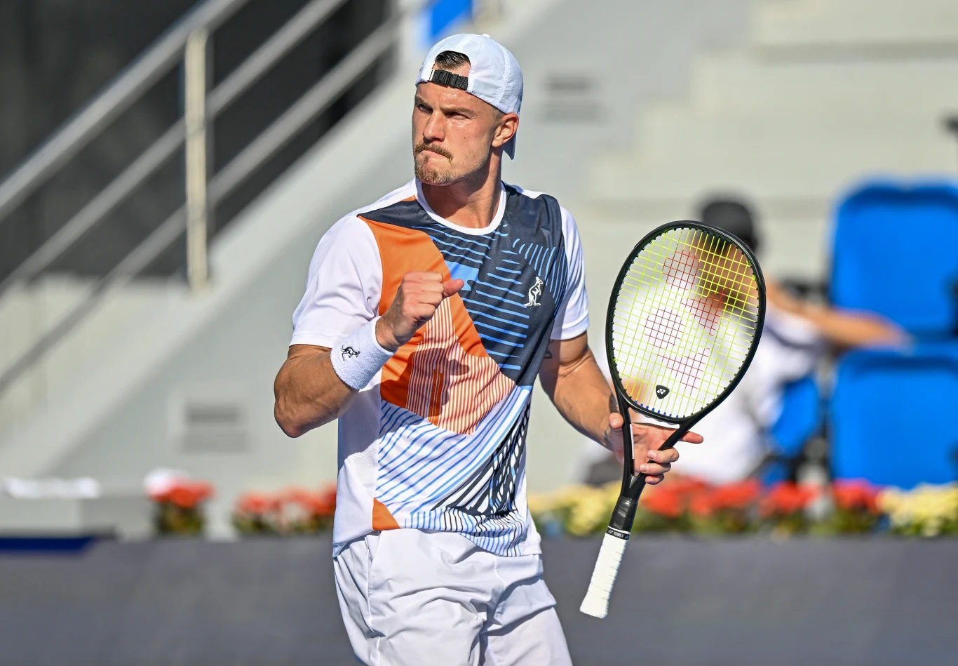 Marton Fucsovics Tenisista w sportowym stroju i czapce z daszkiem, trzymający rakietę, wykonuje gest triumfu na korcie tenisowym podczas meczu.