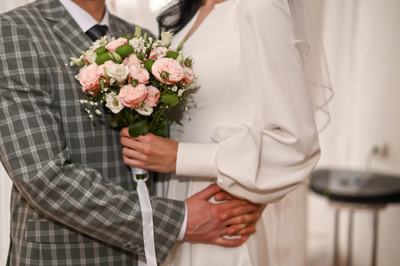 Para młoda stojąca blisko siebie, panna młoda ubrana w białą suknię i trzymająca bukiet różowych i białych kwiatów, pan młody w szarej, kraciastej marynarce obejmuje ją w talii.