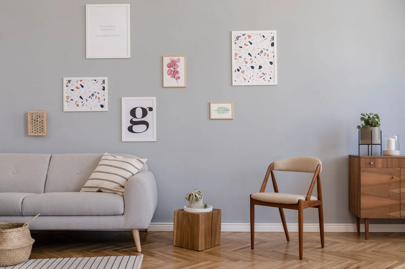 Szara sofa z dekoracyjną poduszką, drewniane krzesło i komoda na tle szarej ściany ozdobionej minimalistycznymi obrazkami oraz rośliną doniczkową na meblu.