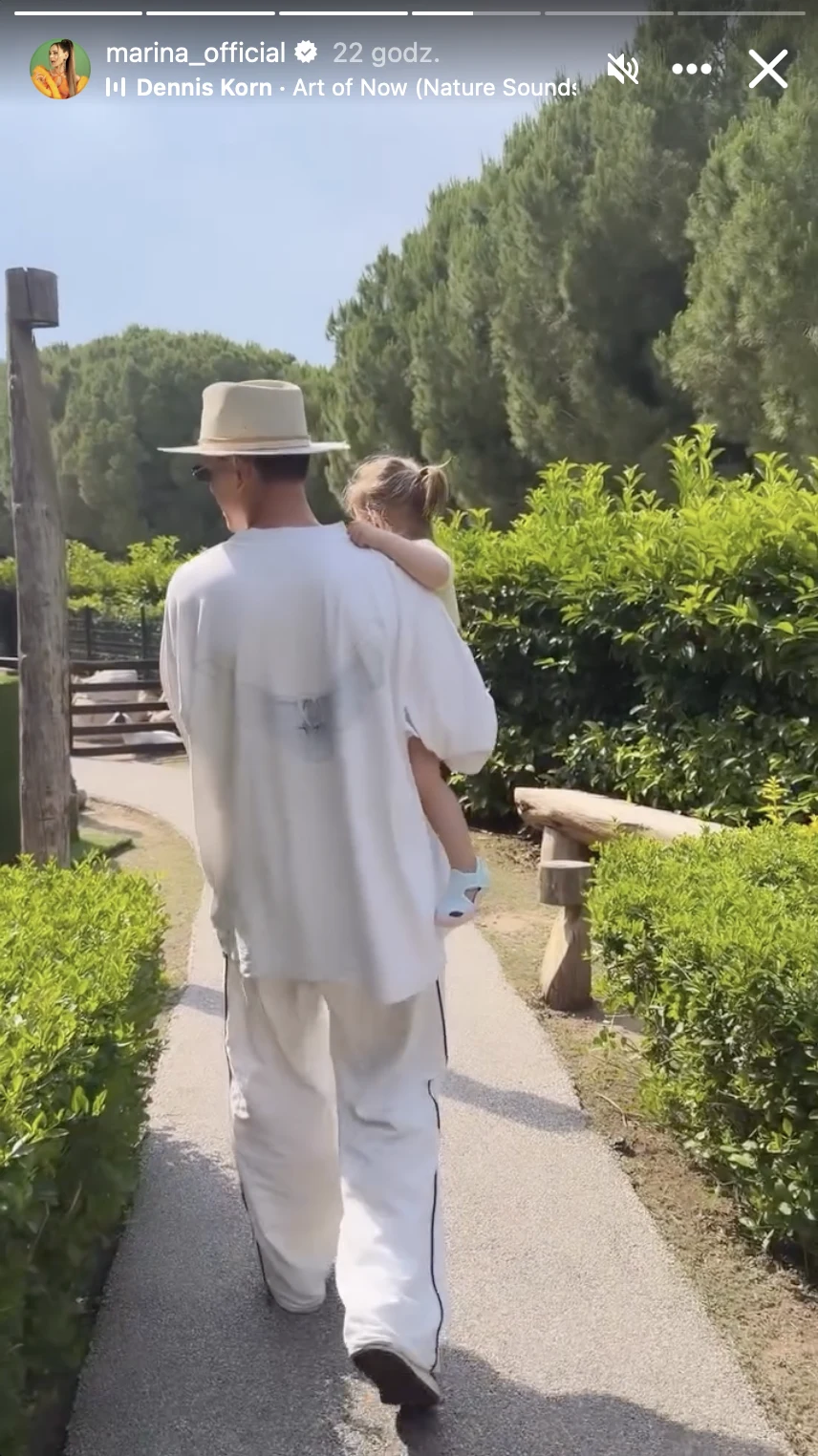 Mężczyzna w białym stroju i kapeluszu niesie małe dziecko, spacerując słoneczną ścieżką otoczoną zielonymi krzewami i wysokimi drzewami.