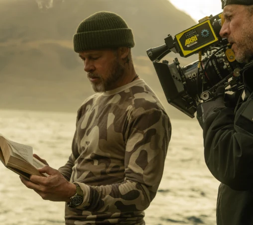 Brad Pitt na planie "Heart of the Beast" Mężczyzna w czapce i bluzie moro stoi nad wodą, trzymając otwartą książkę, obok niego operator filmowy nagrywa scenę profesjonalną kamerą.