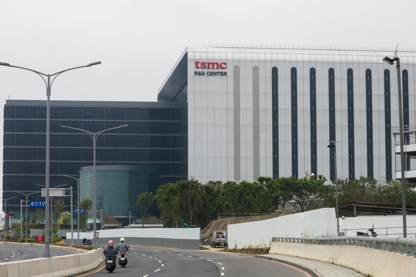Centrum Badawczo-Rozwojowe tajwańskiej firmy TSMC Nowoczesny budynek centrum badawczo-rozwojowego TSMC z dużym logo firmy, szeroka droga z zakrętem i dwoma jadącymi skuterami, otoczenie z zielenią i niskim ogrodzeniem.