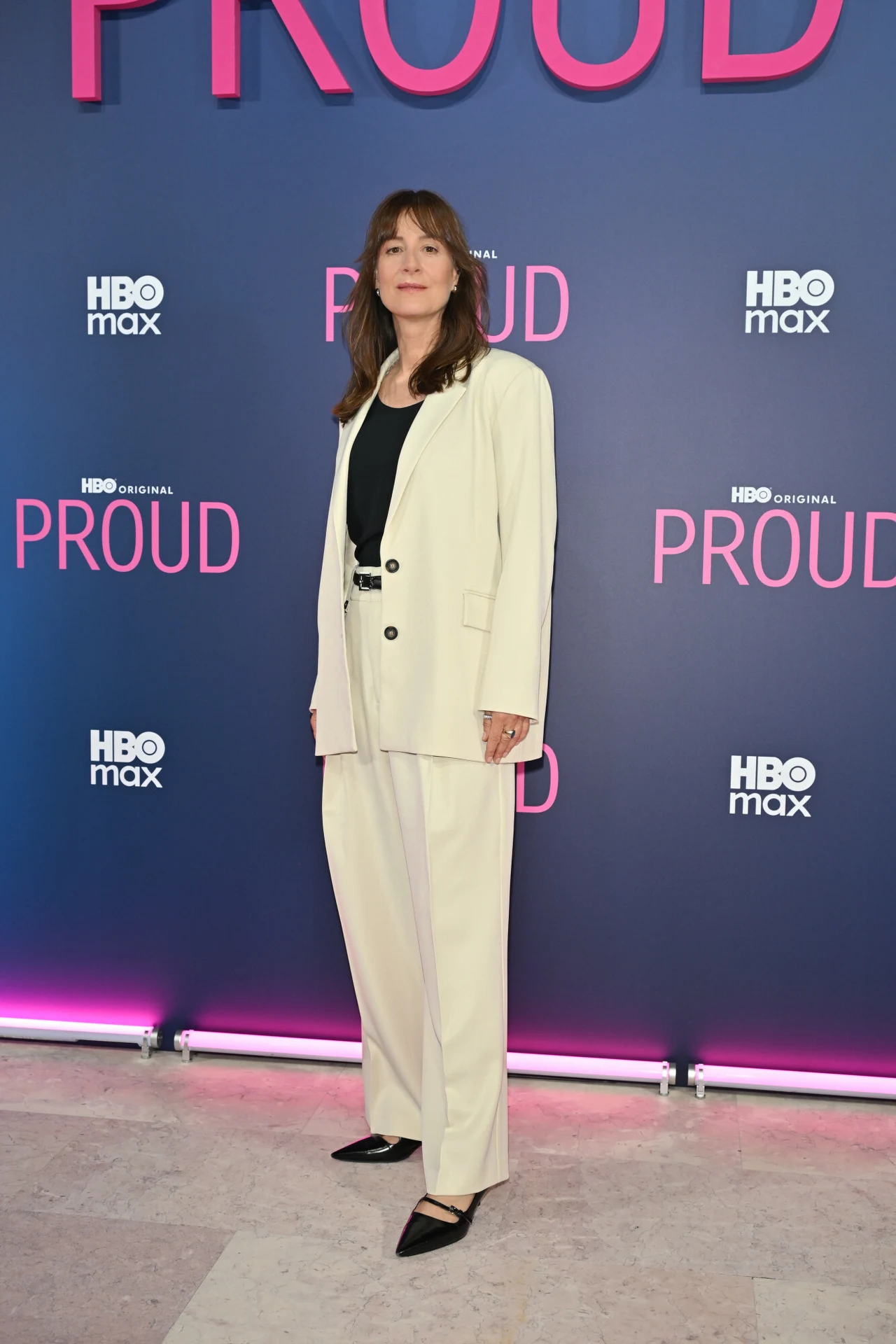 Maja Ostaszewska w białym garniturze na premierze serialu. Klasa i elegancja Kobieta w kremowym garniturze i czarnym topie pozuje na tle ścianki promującej produkcję Proud HBO Max, ubrana w eleganckie czarne buty na niskim obcasie.