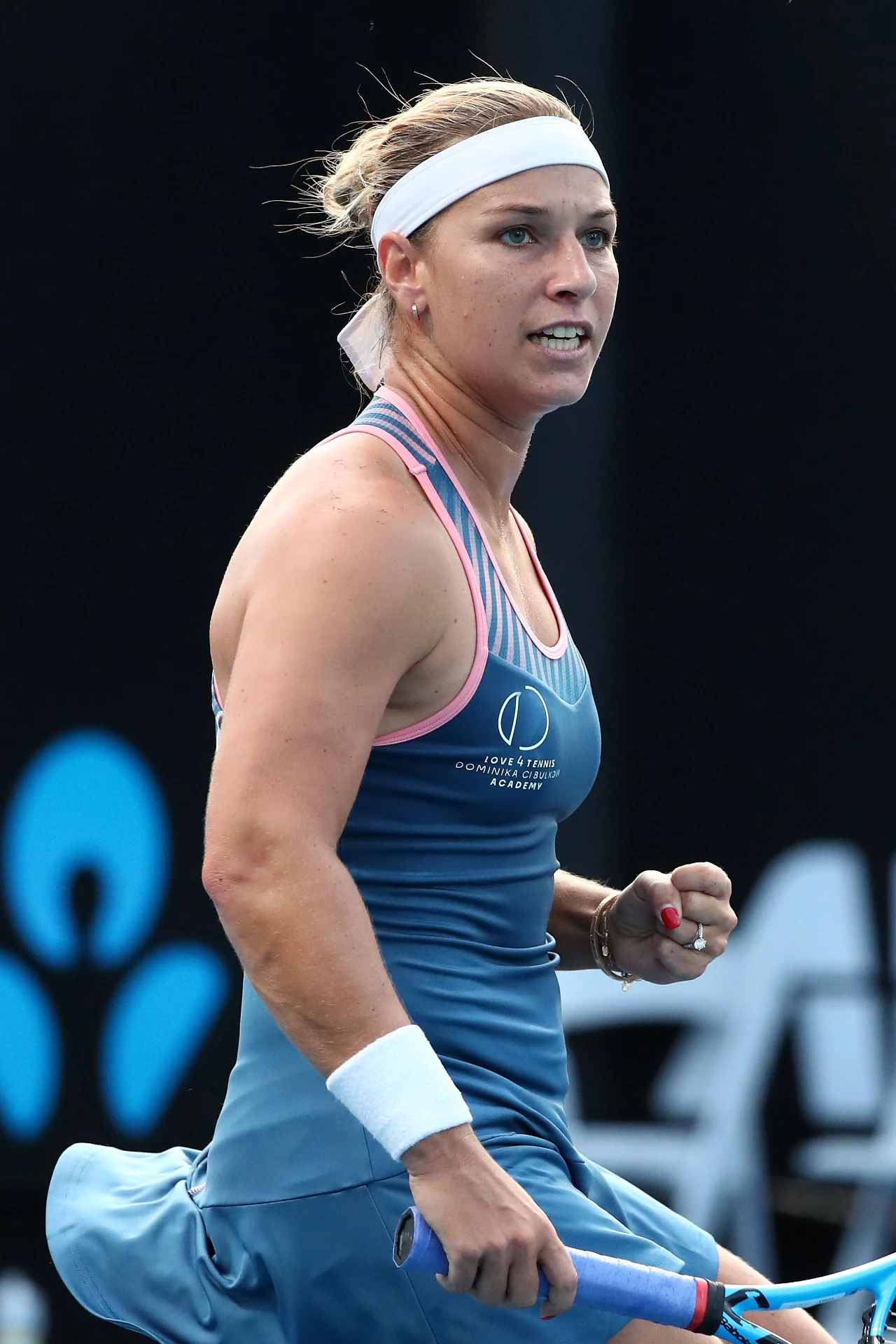 Tenisistka w niebieskiej sukience sportowej i białej opasce na głowie świętuje zdobycie punktu, ściskając pięść z wyrazem determinacji na twarzy.