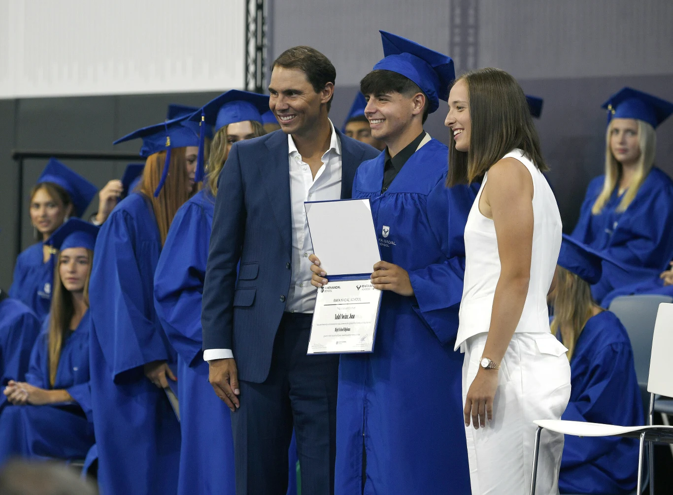 Absolwentka w niebieskiej todze i birecie pozuje z dyplomem obok elegancko ubranego mężczyzny i kobiety w białym kombinezonie, otoczona innymi osobami w niebieskich togach siedzącymi w tle.