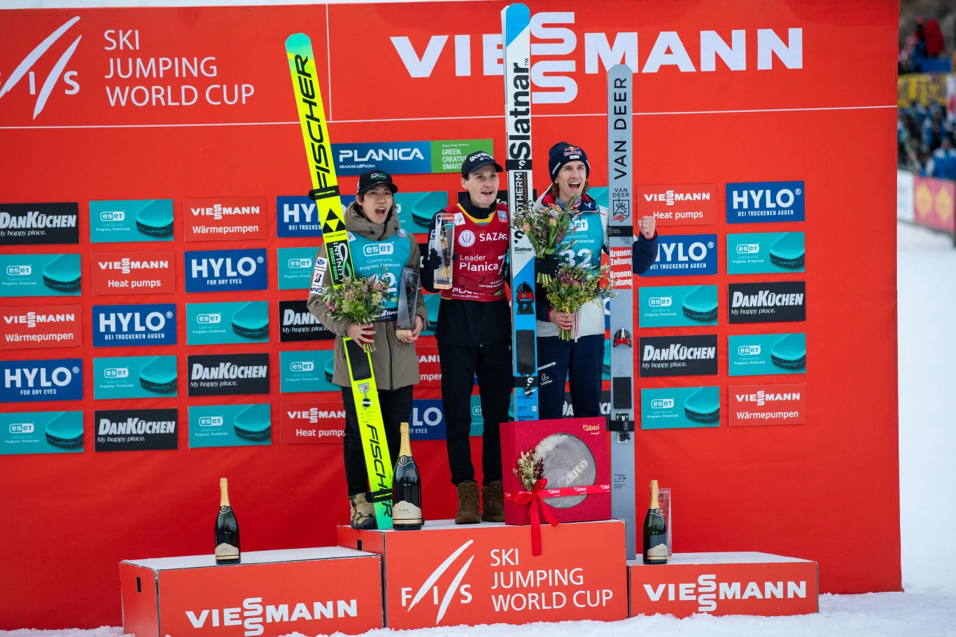 Trzech skoczków narciarskich stojących na podium z nartami w rękach, ubranych w kombinezony sportowe i czapki, trzymających wiązanki kwiatów na tle ścianki sponsorskiej podczas zawodów Pucharu Świata w skokach narciarskich.