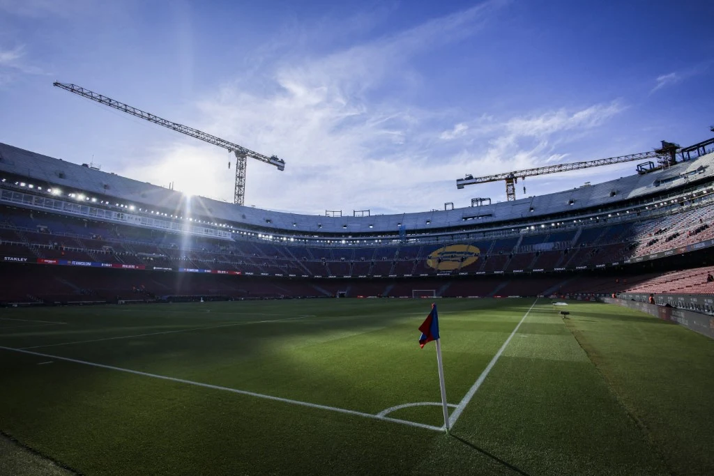 Pusta murawa stadionu piłkarskiego otoczona trybunami oraz dwoma wysokimi dźwigami budowlanymi na tle błękitnego nieba, w jednym z rogów boiska stoi chorągiewka z niebiesko-czerwonymi barwami.