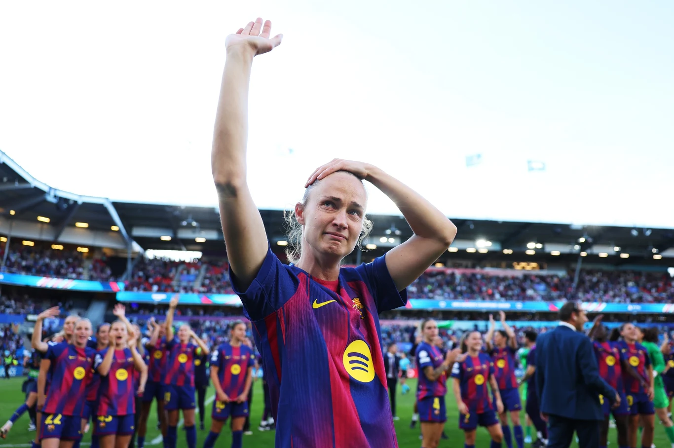 Piłkarka w stroju FC Barcelony z uniesioną ręką na tle grupy drużyny świętującej na stadionie.