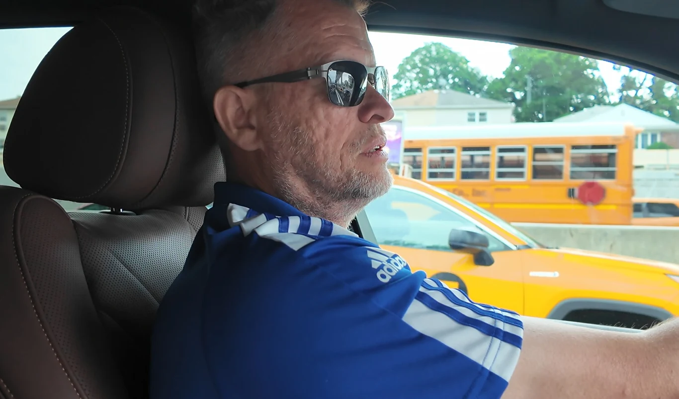 Mężczyzna w okularach przeciwsłonecznych prowadzi samochód, ubrany w niebieską koszulkę sportową, za oknem widoczny żółty autobus szkolny i samochód osobowy.