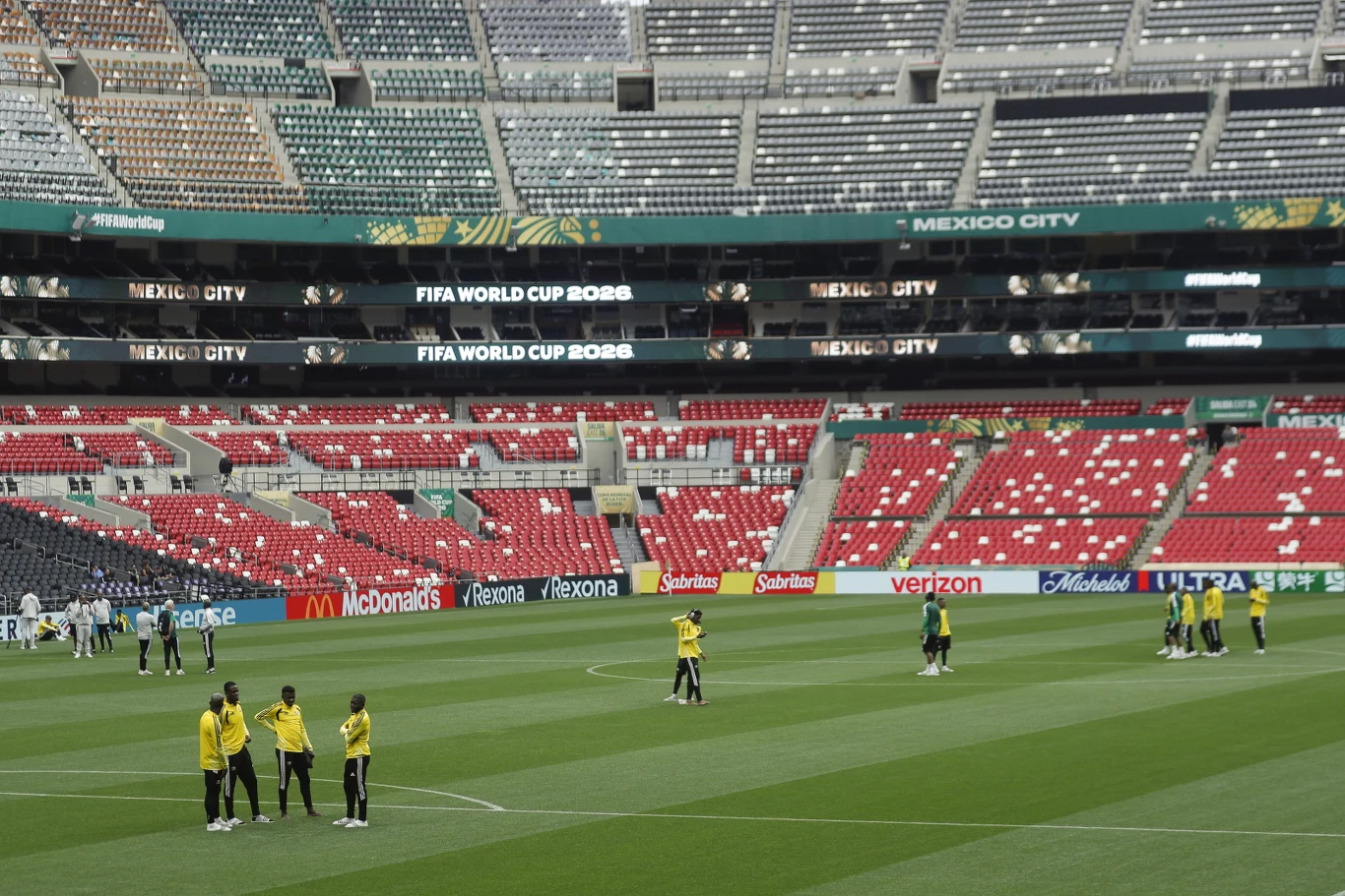 Piłkarze w żółtych koszulkach rozgrzewają się na zielonej murawie stadionu otoczonego rzędami pustych czerwonych i szarych krzesełek, z wyraźnymi tablicami informującymi o Mistrzostwach Świata FIFA 2026 oraz lokalizacją Mexico City.