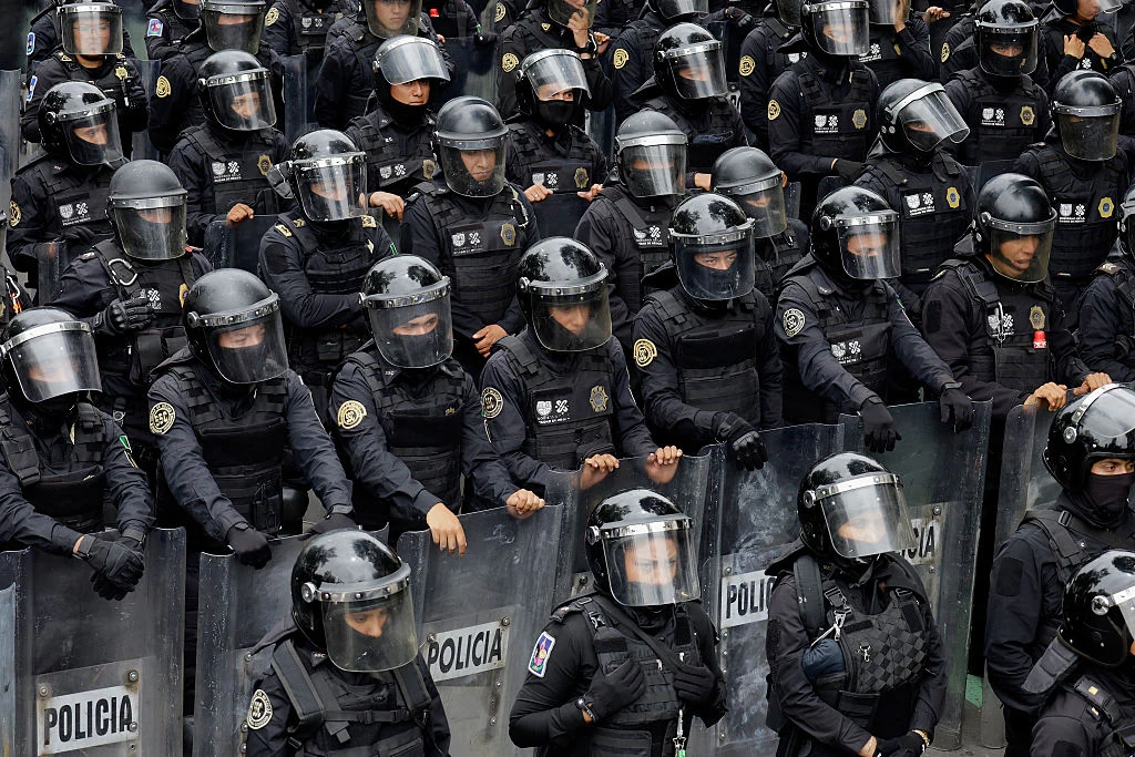 Zgrupowanie funkcjonariuszy policji w pełnym umundurowaniu ochronnym, wyposażonych w kaski, przyłbice i przezroczyste tarcze, tworzących zwartą linię zabezpieczającą teren.