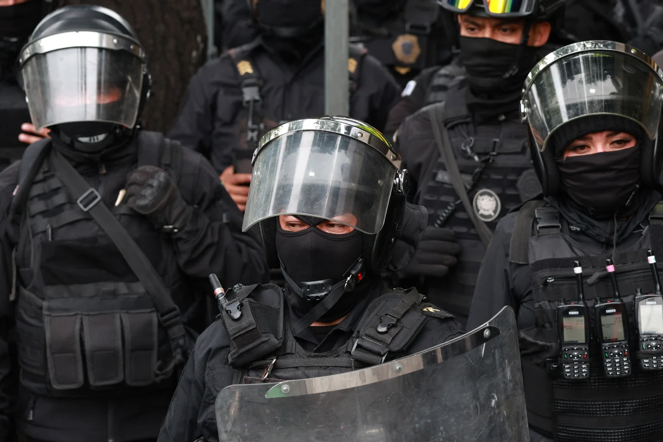 Grupa policjantów w pełnym umundurowaniu ochronnym z hełmami i przyłbicami, wyposażeni w tarcze oraz radiotelefony, stoi w zwartym szyku.