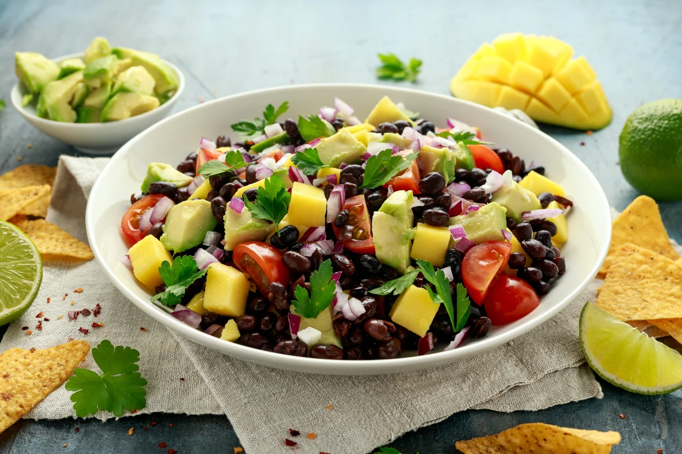 Czarna fasola świetnie komponuje się w sałatkach z kukurydzą, awokado oraz kolendrą. Sałatka z czarnej fasoli z dodatkiem awokado, mango, czerwonej cebuli, pomidorków koktajlowych i świeżej kolendry, podana w misce, otoczona kawałkami limonki, nachosami i dodatkami w formie pokrojonego awokado oraz mango.