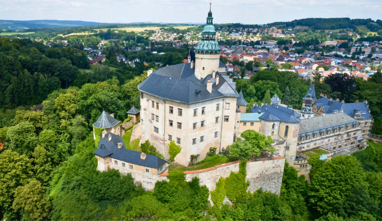 Zamek w stylu renesansowym usytuowany na wzgórzu, otoczony gęstą zielenią, z rozległym widokiem na miasteczko i pagórkowaty krajobraz w tle.