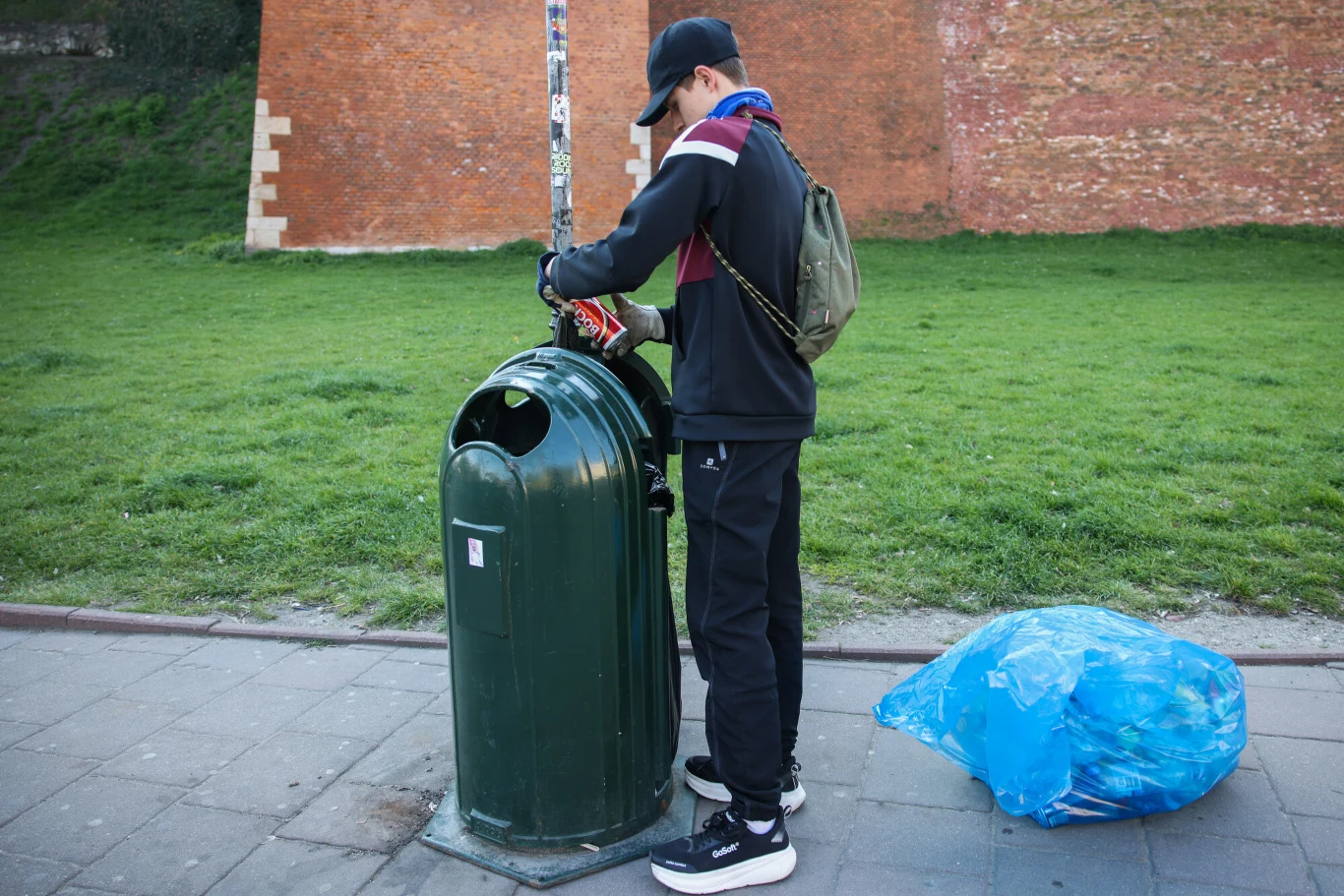Młody mężczyzna w czapce i plecaku wyrzuca śmieci do dużego zielonego kosza na ulicy, obok niego leży niebieski worek foliowy pełen odpadów.