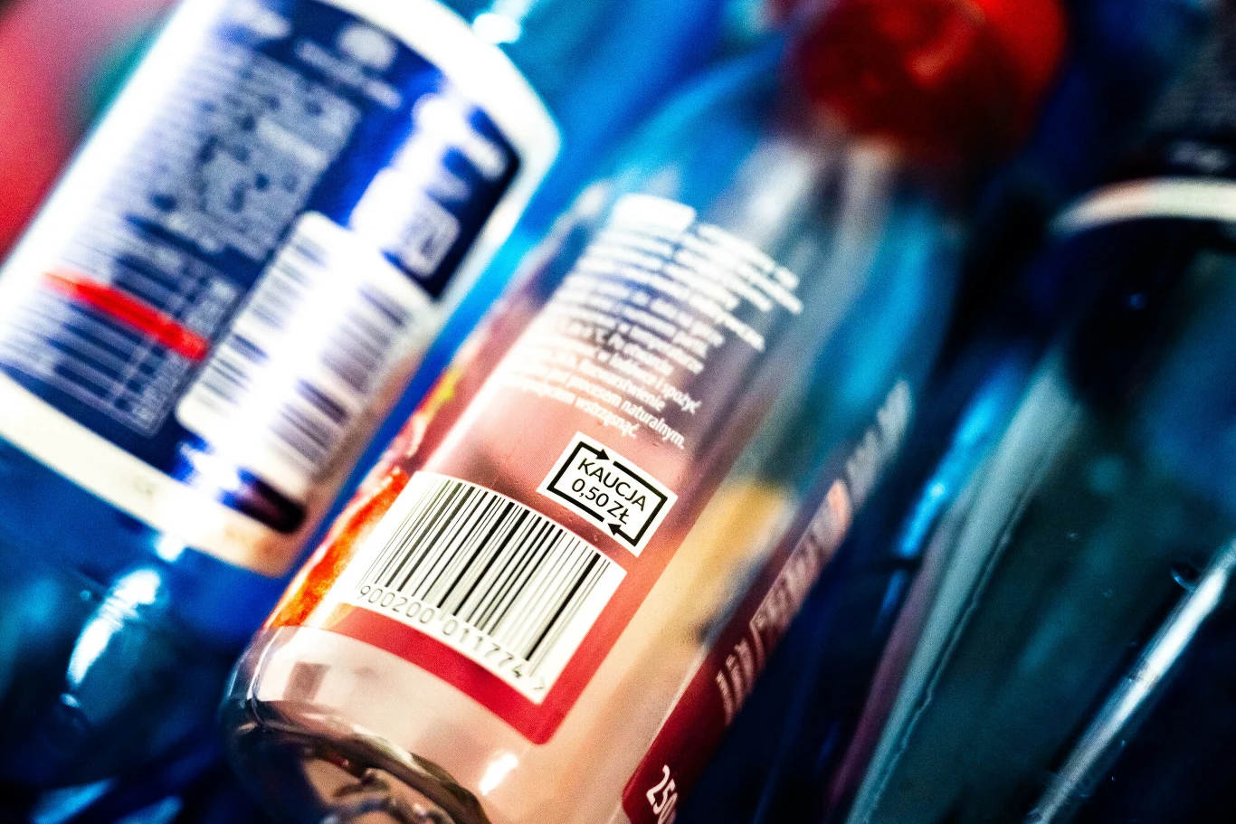 Zbliżenie na plastikowe butelki, z których jedna posiada widoczną etykietę z oznaczeniem kaucji oraz kodem kreskowym.