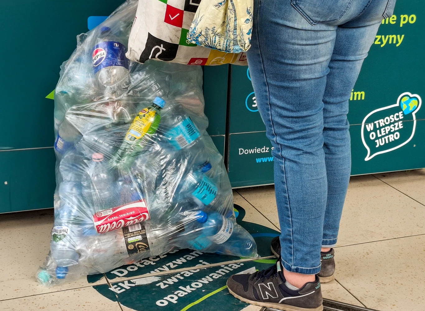 Woreczek wypełniony zużytymi plastikowymi butelkami stoi przy automacie do recyklingu, obok osoba w dżinsach i sportowych butach, trzymająca w ręku torbę na zakupy.