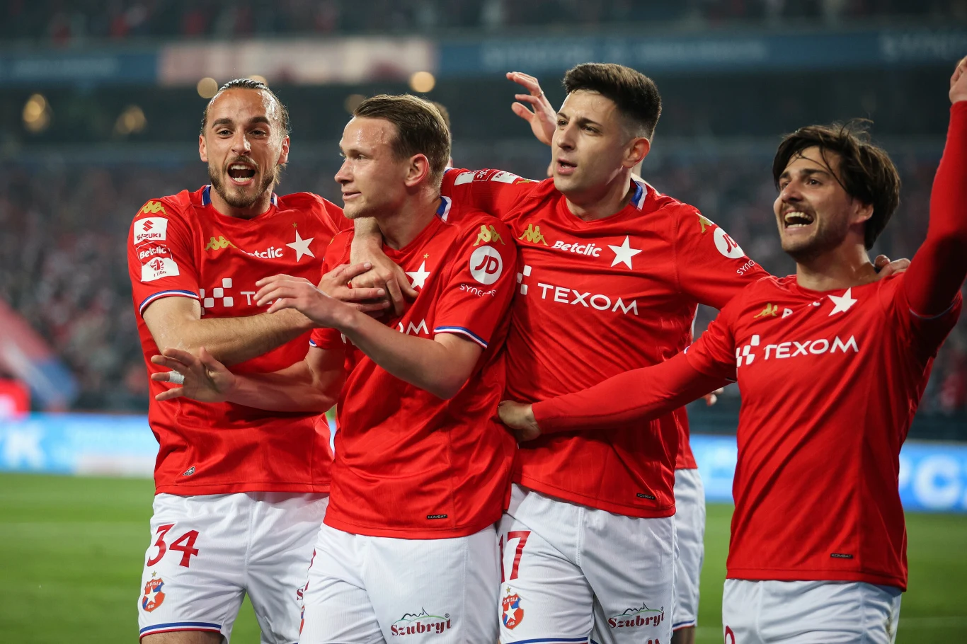 Wisła Kraków Czterech piłkarzy w czerwonych koszulkach świętuje zdobycie gola na murawie stadionu, obejmując się i wyrażając radość.