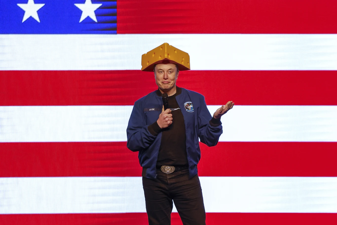 Elon Musk w niebieskiej kurtce i czapce przypominającej ser, na tle amerykańskiej flagi, z mikrofonem.