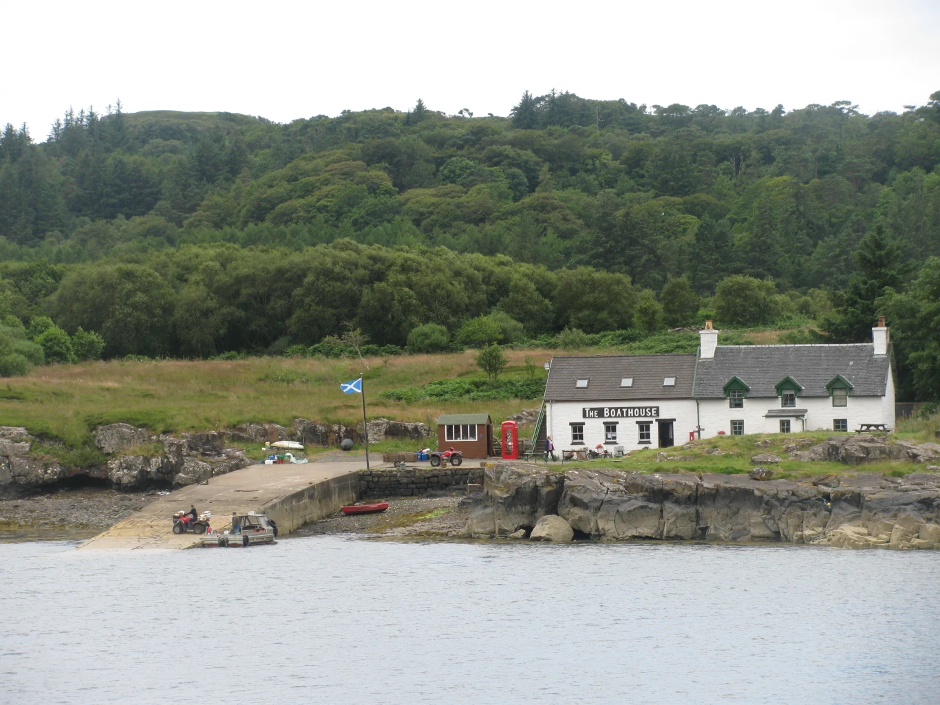 Biały budynek z napisem „The Old Forge”, szkocka flaga na maszcie, mały port z zacumowanymi łodziami i gęsty, zielony las w tle.
