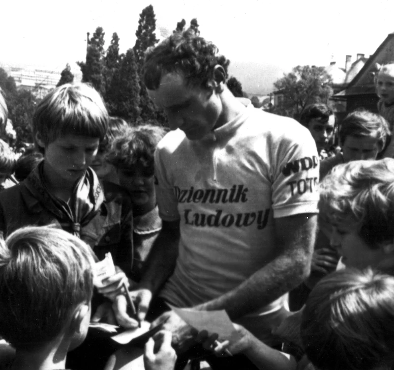 Mężczyzna w sportowej koszulce z napisem „Dziennik Ludowy” rozdaje autografy grupie dzieci skupionych wokół niego.