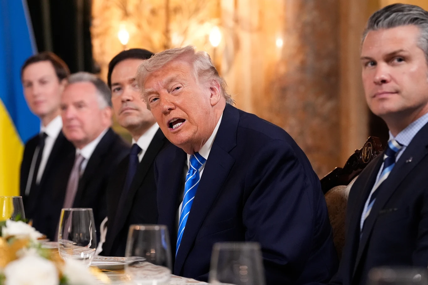 Prezydent Stanów Zjednoczonych Donald Trump w otoczeniu swoich najbliższych współpracowników. Od lewej: Jareda Kushnera, Steve'a Witkoffa, Marco Rubio i Pete'a Hegsetha Pięciu elegancko ubranych mężczyzn siedzących przy stole, po lewej stronie widoczny fragment flagi Ukrainy, centralnie postać z jasnymi włosami mówi podczas spotkania.