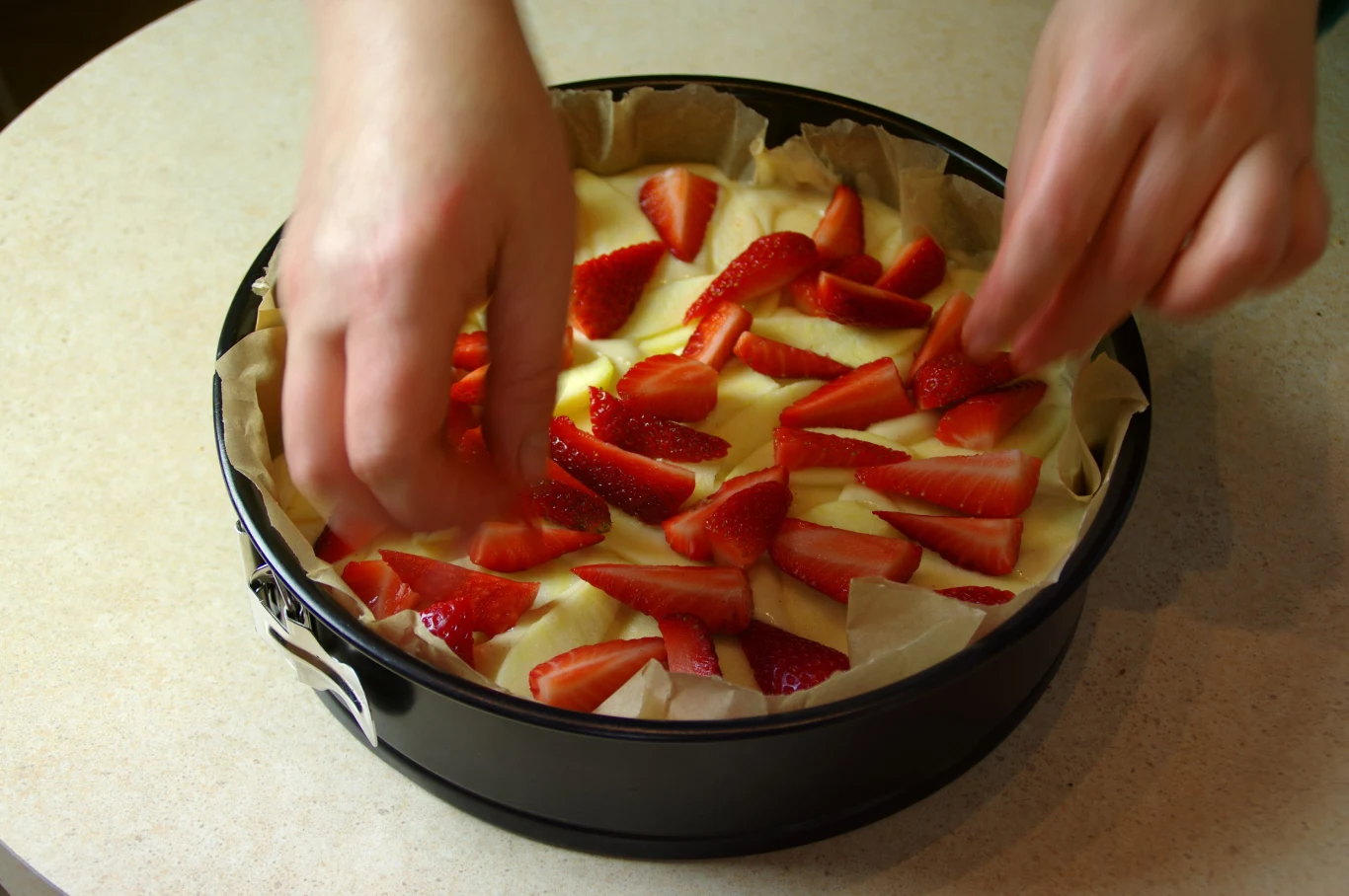 Preparing yeast cake with apple and strawberry to baking. Fresh raw homemade fruit pastry. Ręce układają plasterki truskawek na jasnym cieście w okrągłej formie wyłożonej papierem do pieczenia.