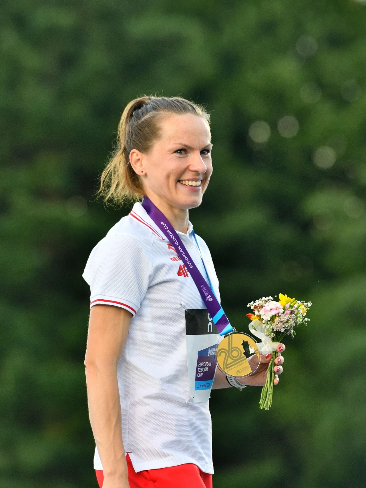 Uśmiechnięta kobieta w biało-czerwonym stroju sportowym z medalem na szyi i bukietem kwiatów w dłoni.