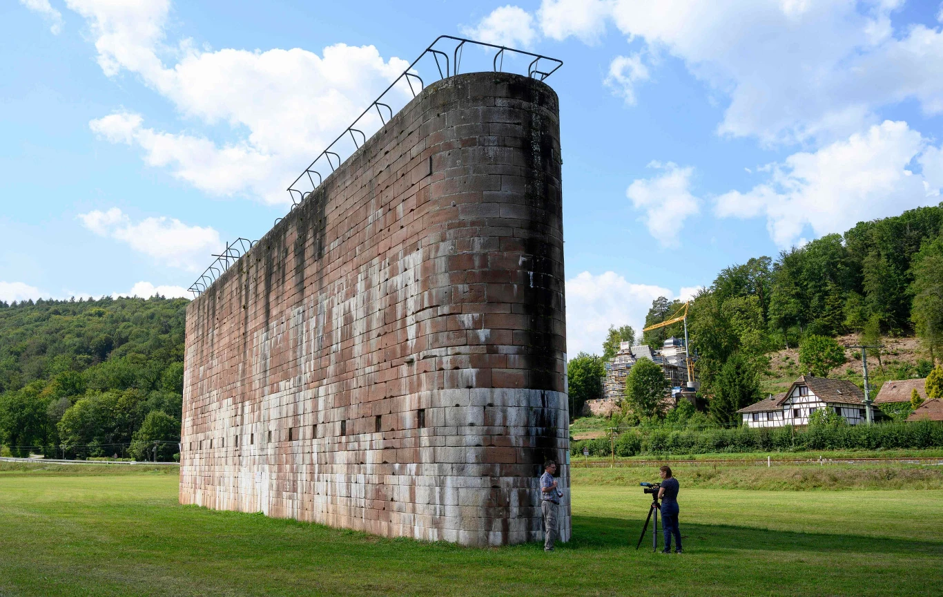 Masynowy, masywny fragment historycznego muru z czerwonego kamienia z metalową barierką na górze, przed którym stoi dwoje ludzi z aparatem, otoczony otwartą zieloną przestrzenią i drzewami.