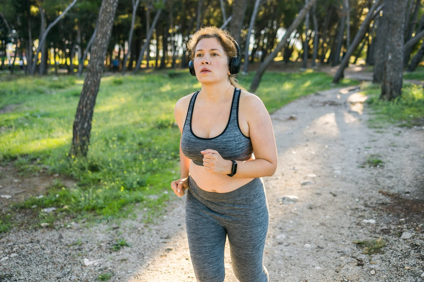 Intensywność treningów powinna być dopasowana do kondycji, sprawności, jak również masy ciała Kobieta w sportowym stroju biegnie leśną ścieżką, na głowie ma słuchawki bezprzewodowe.