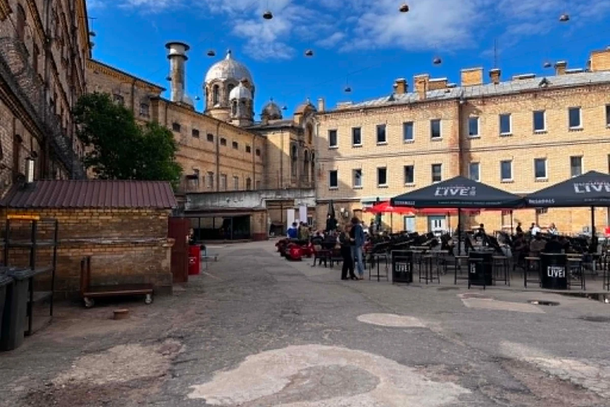 Zabytkowy dziedziniec otoczony wysokimi, ceglastymi budynkami, w którego centralnej części ustawiono stoliki i czarne parasole, a w tle widać kopułę i kilka osób korzystających z przestrzeni kawiarnianej.