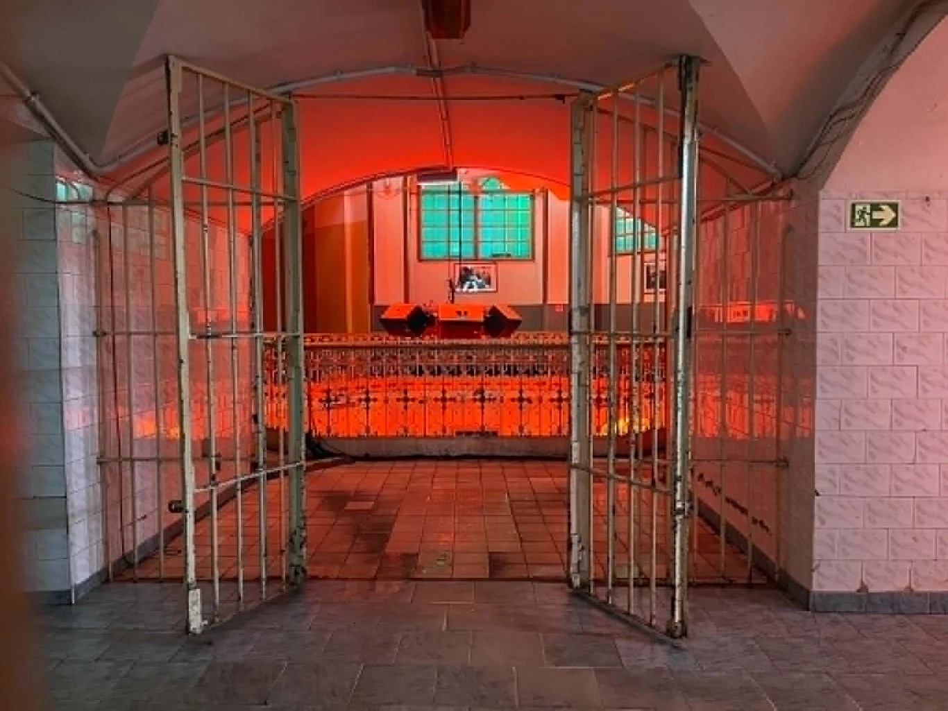 Otwarta brama z metalowych krat prowadzi do pomieszczenia oświetlonego intensywnym czerwonym światłem, w tle widoczne są ozdobne balustrady oraz jasnozielone okna.
