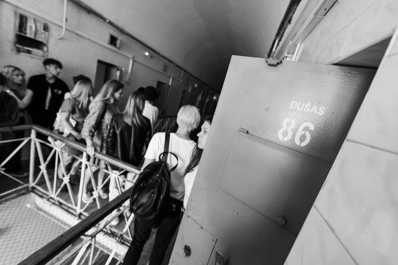 Grupa ludzi stojących na korytarzu przy otwartych drzwiach z numerem 86 i napisem DUŠAS, częściowo wchodzących do wnętrza lub oczekujących przed wejściem.