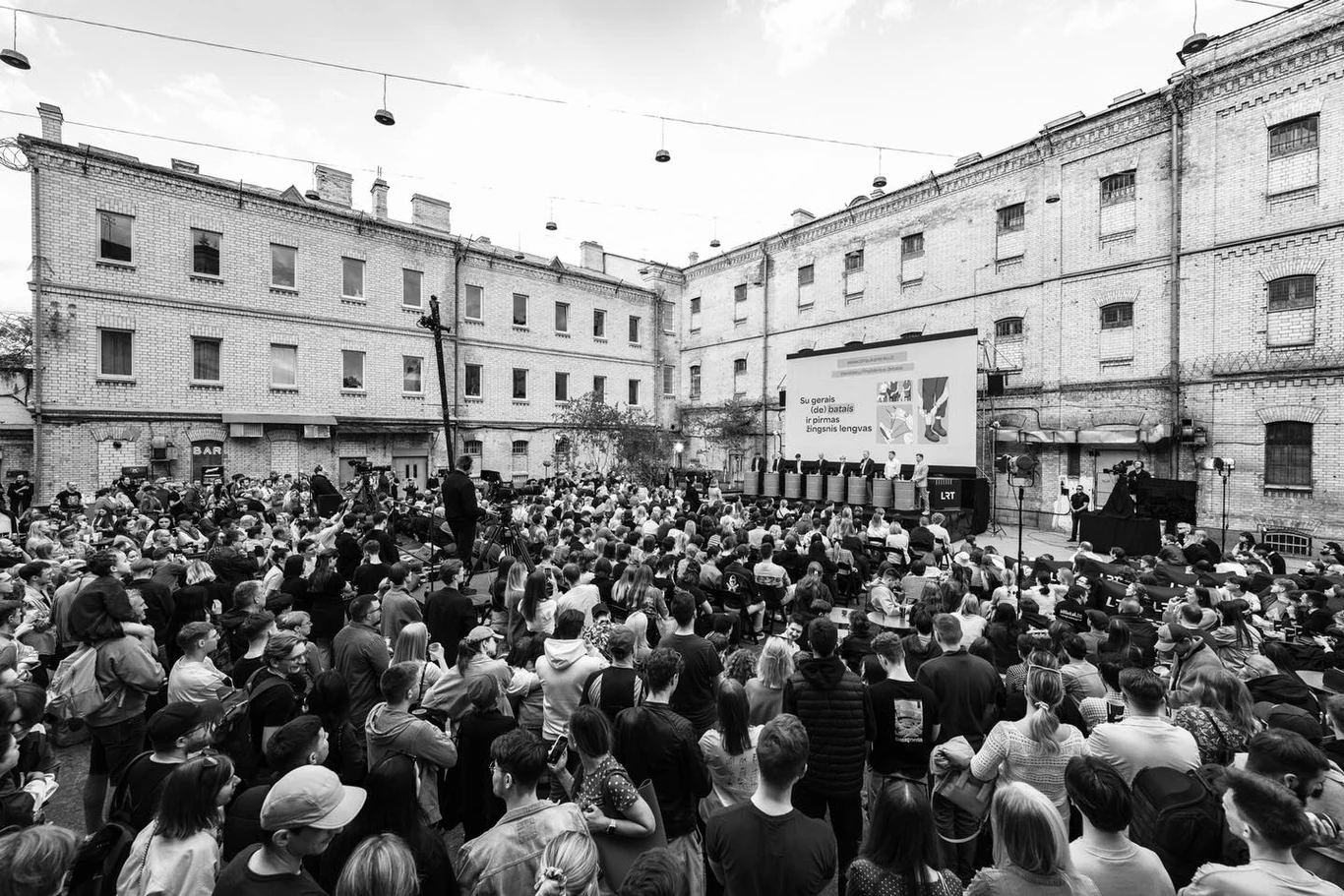 Duży tłum ludzi zgromadzony na dziedzińcu otoczonym starymi ceglastymi budynkami, skierowany w stronę sceny z dużym ekranem i osobami prowadzącymi wydarzenie.