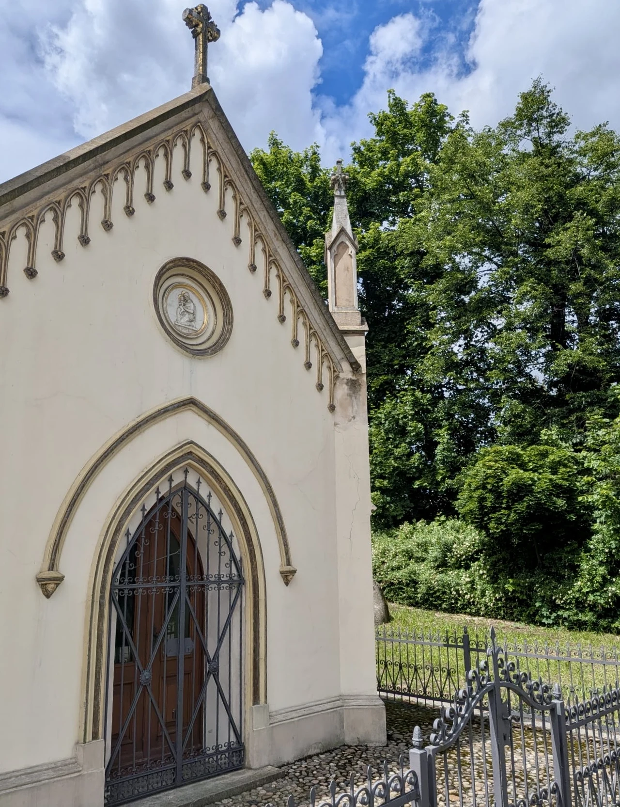 Kapliczka św. Jana Nepomucena w Łabędach - jeden ze śladów pozostałych po rodzie von Welczek w Łabędach Gotycka kaplica z ozdobnym portalem i zamkniętą kratą, udekorowana medalionem z wizerunkiem oraz krzyżem na szczycie, otoczona metalowym ogrodzeniem; w tle gęste drzewa pod błękitnym niebem z chmurami.