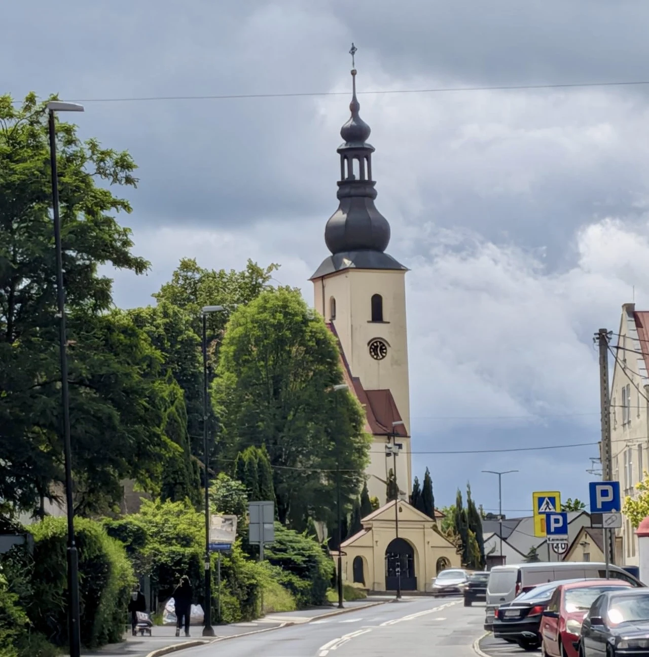 Kościół Wniebowzięcia NMP w Gliwicach Łabędach Kościół z wysoką wieżą i charakterystyczną ciemną kopułą, otoczony zielenią i budynkami, przy głównej ulicy z zaparkowanymi samochodami oraz oznakowanymi miejscami parkingowymi.
