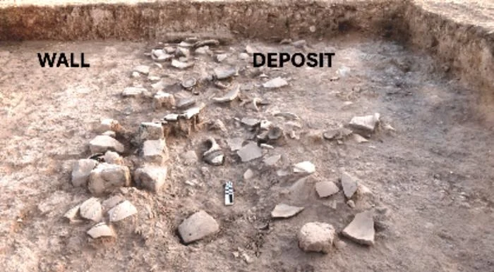 Fragmenty kamieni i ceramiki rozrzucone w wykopie archeologicznym, z wyraźnie zaznaczonymi strefami muru i nagromadzenia znalezisk.