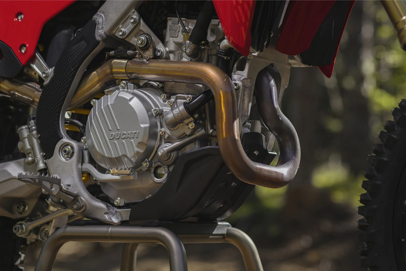 silnik motocykla Ducati z wyraźnie widocznym wydechem oraz elementami ramy i osłoną silnika, podparty na metalowym stojaku