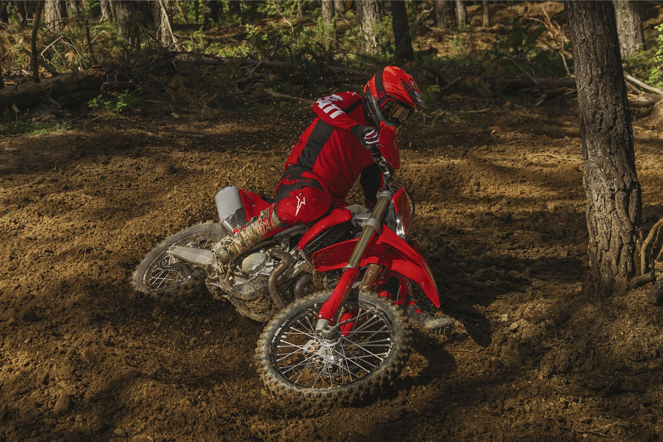 Motocyklista w czerwonym kombinezonie pokonuje zakręt na off-roadowym motocyklu jadąc przez leśny teren o miękkim, błotnistym podłożu.