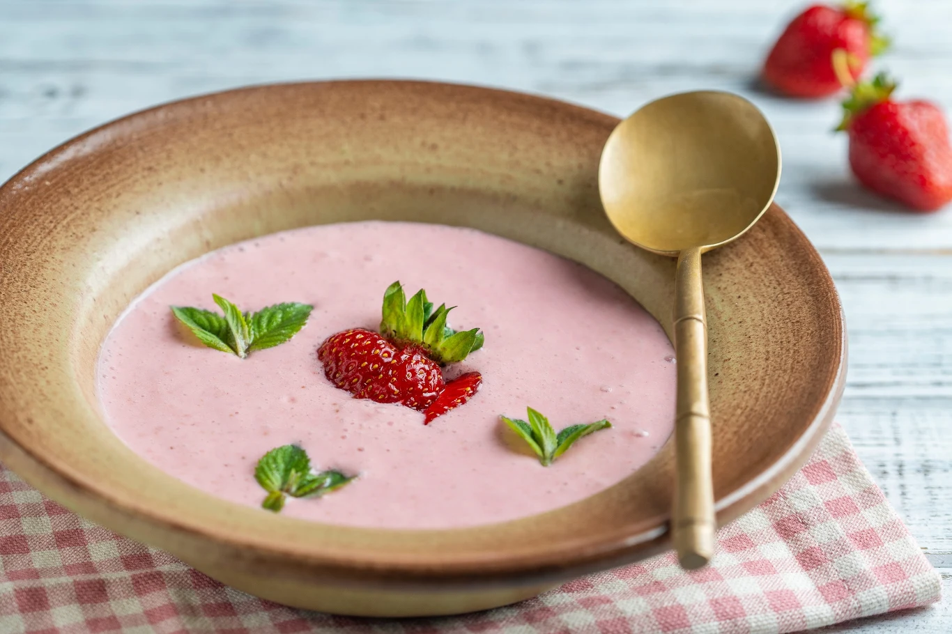 Zupa truskawkowa Truskawkowa zupa krem ozdobiona listkami mięty i kawałkami truskawek, podana w głębokim talerzu ze złotą łyżką na lnianej serwetce.