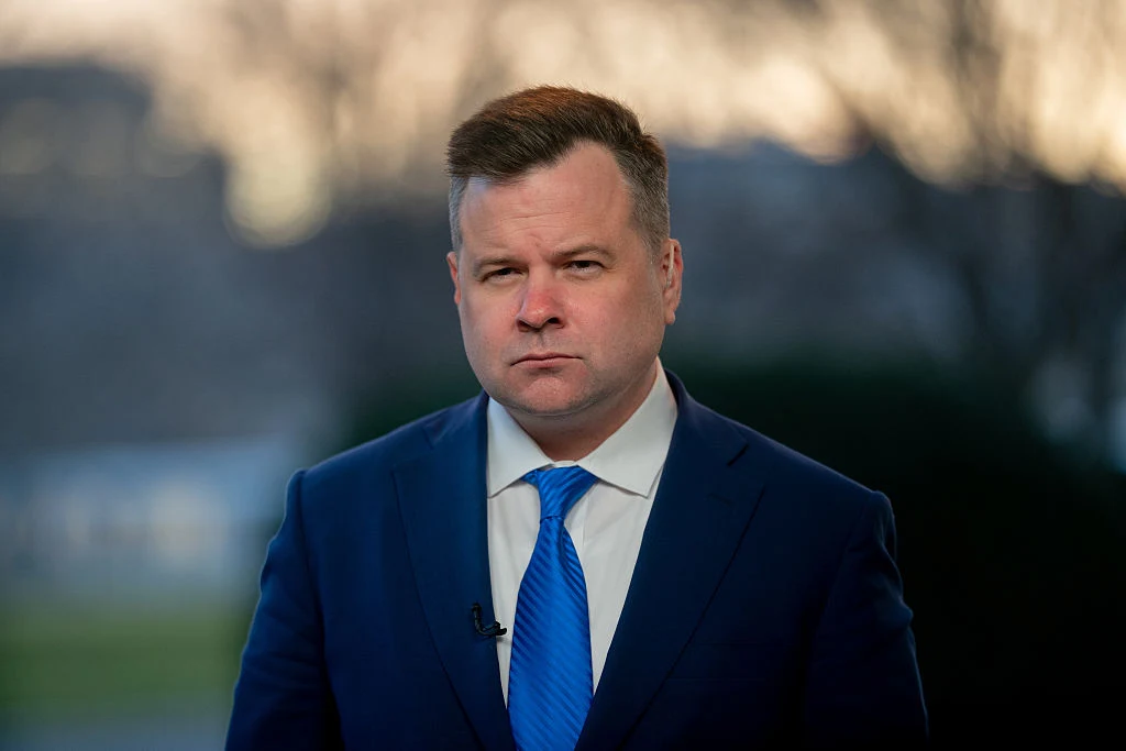 Mężczyzna w granatowym garniturze i niebieskim krawacie stoi na zewnątrz, patrząc poważnie przed siebie.