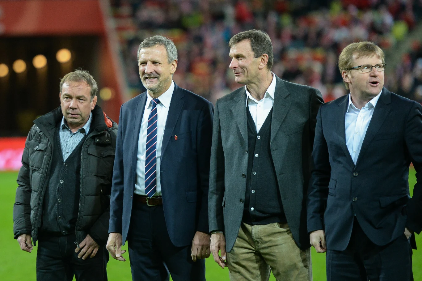 Czterech elegancko ubranych mężczyzn stoi obok siebie na stadionie sportowym, uśmiechając się i kierując wzrok w różne strony, w tle zamazana publiczność na trybunach.