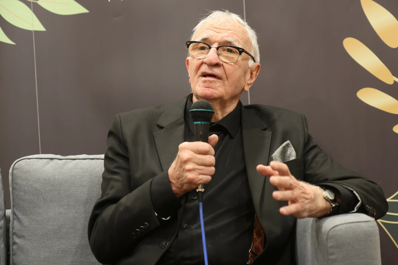 Starszy mężczyzna w czarnym garniturze i okularach siedzi na sofie, trzyma mikrofon i gestykuluje podczas wypowiedzi.
